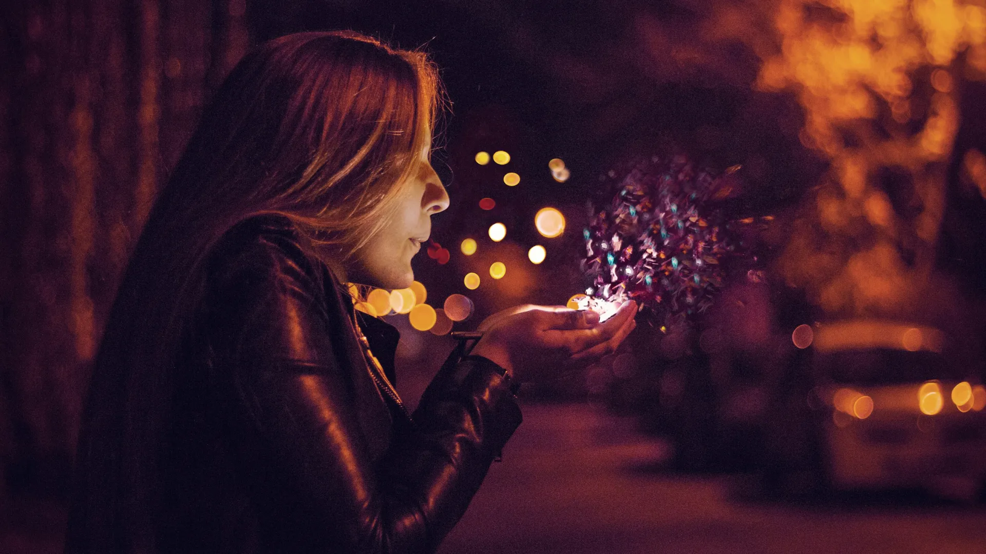 Young woman in leather jacket blowing colorful confetti from hands on a warmly lit street at night.