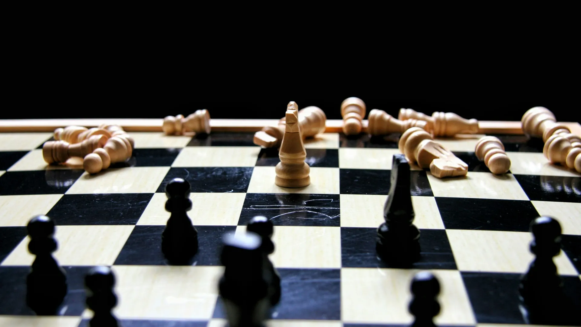 A chessboard with scattered white pieces and standing black pieces in focus against a dark background.