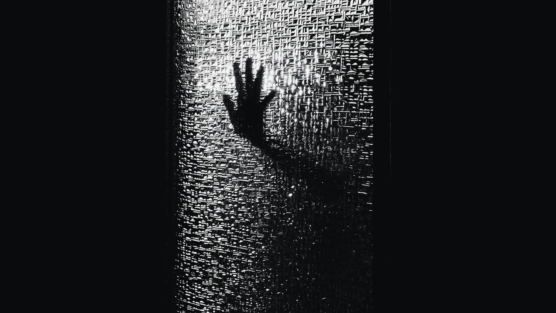 Silhouette of a hand pressed against textured glass in a dark room with light shining through.