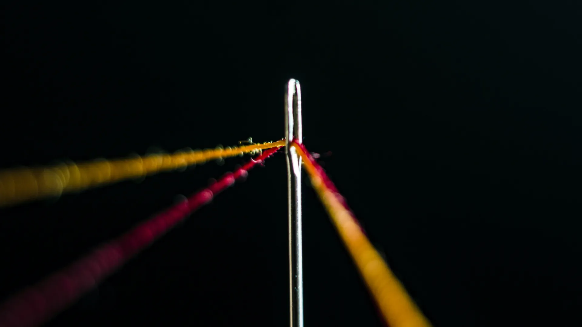 Close-up of a needle eye threaded with vibrant yellow and pink threads against a black background.