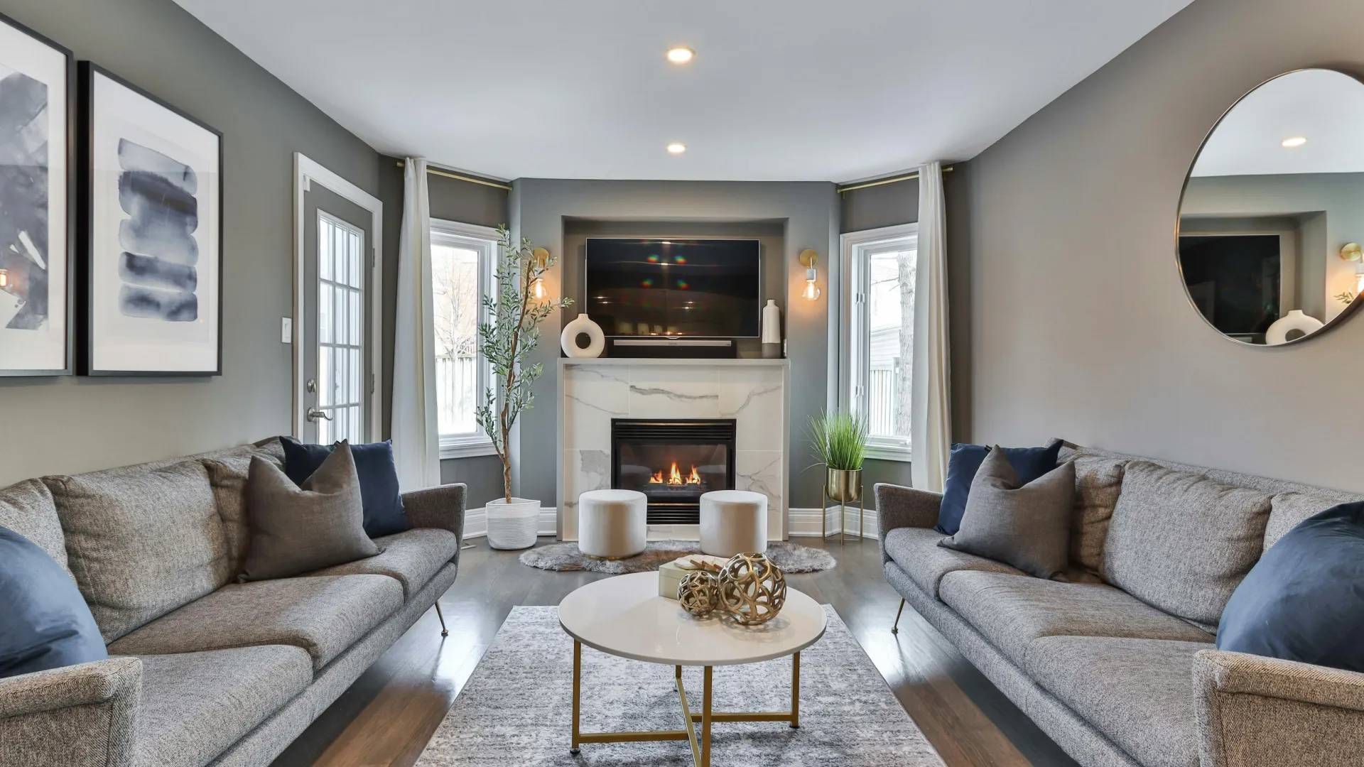 Modern living room with gray sofas, fireplace, wall art, round mirror, and a white coffee table on a rug