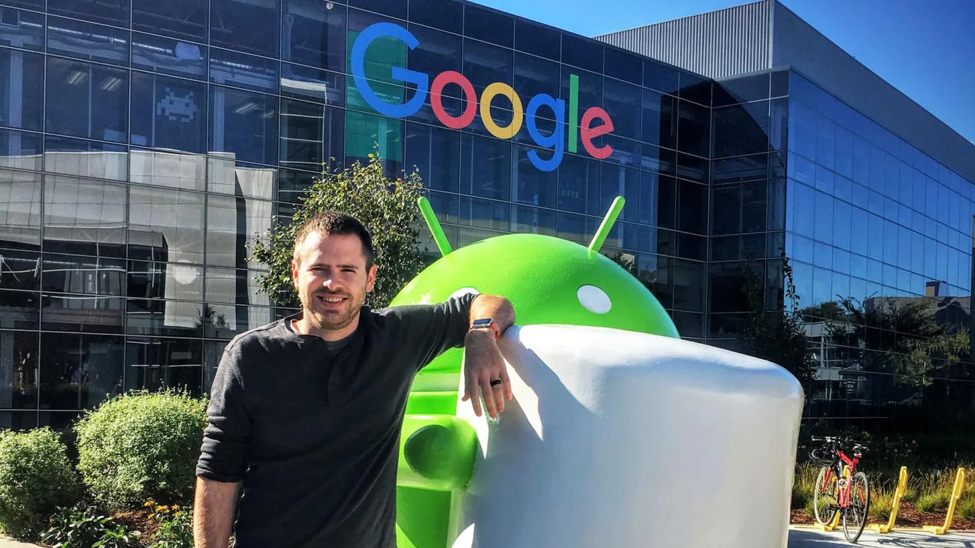 Man posing with a large Android Marshmallow statue outside the Google building with reflective glass windows.