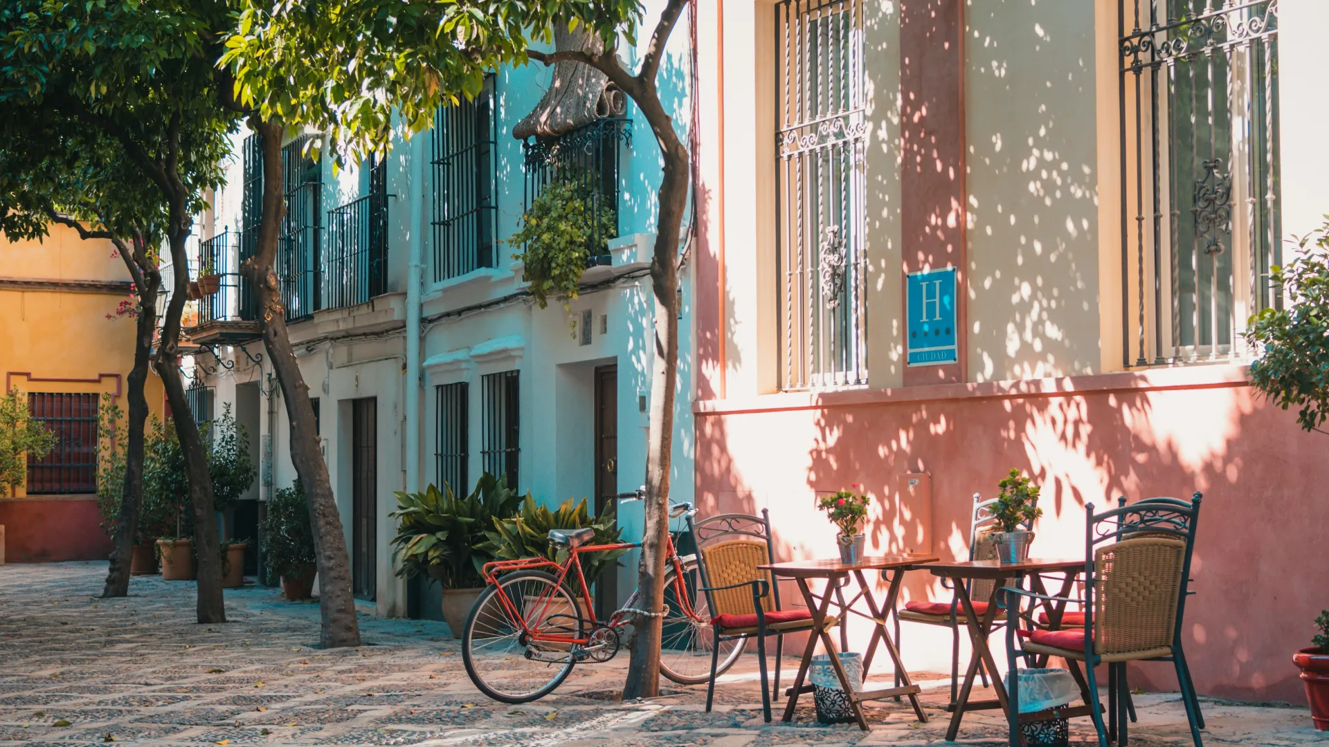 Sunlit outdoor café seating with tables, chairs, a red bicycle, and colorful buildings with trees in a cobblestone street.