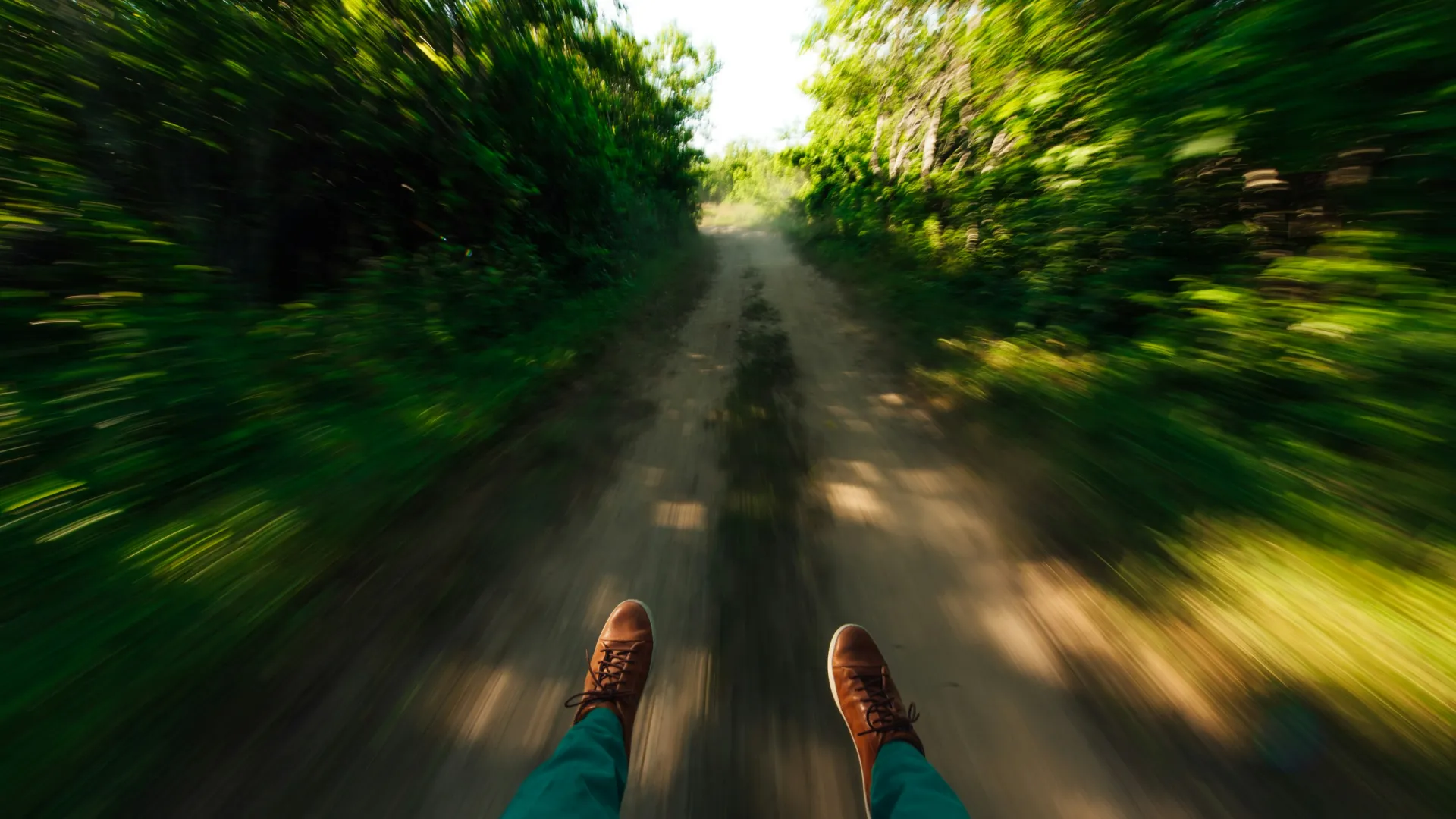 Person wearing brown shoes and green pants moving swiftly down a dirt path surrounded by green trees.