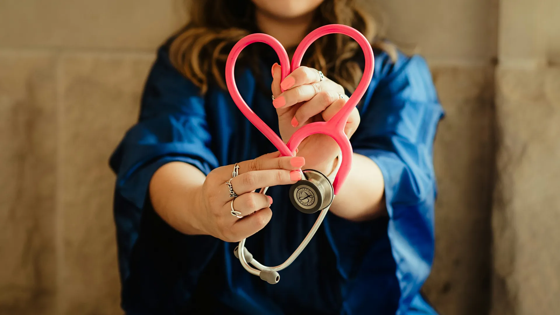 Woman holding a pink stethoscope shaped into a heart symbol with blurred background.