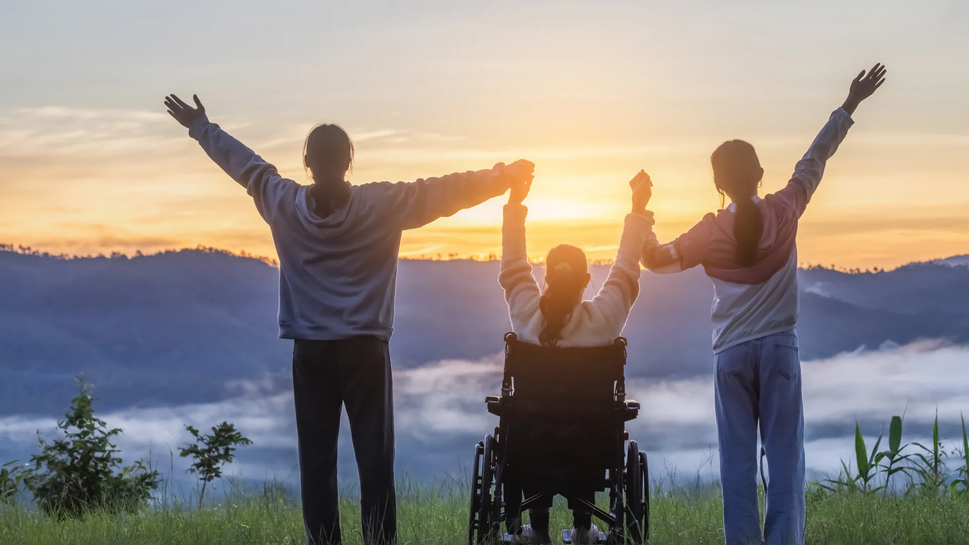 Three people including a person in a wheelchair holding hands and raising arms at sunrise in a scenic mountain field.
