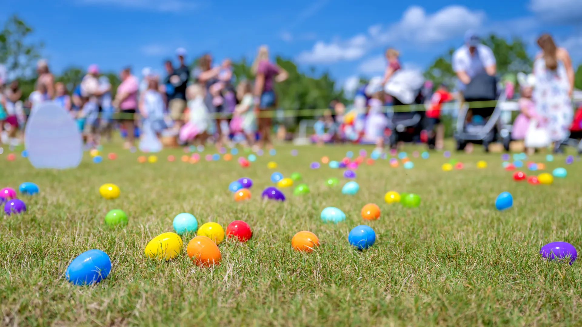 Colorful plastic eggs scattered on grass during a sunny outdoor Easter egg hunt with families in the background