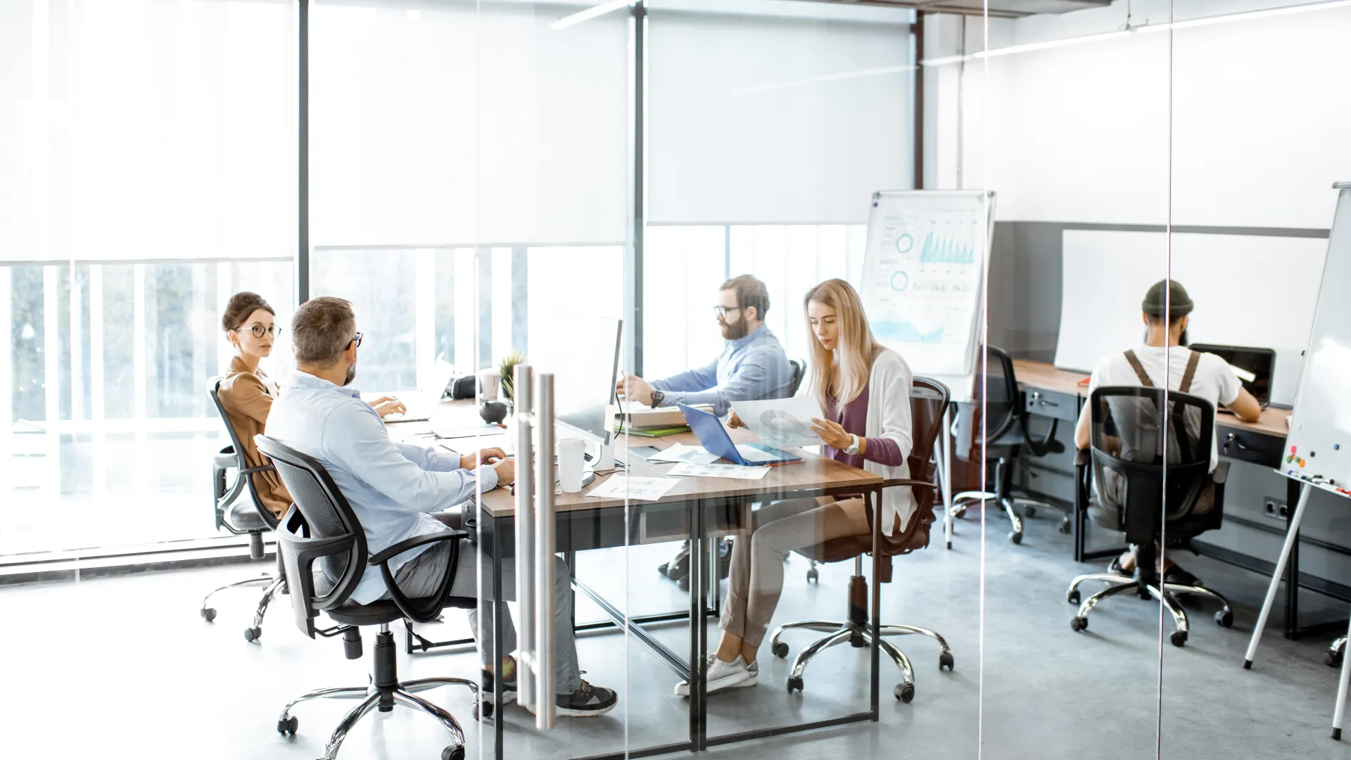 Business team working inside a modern glass office with laptops, documents, and charts in a collaborative meeting.