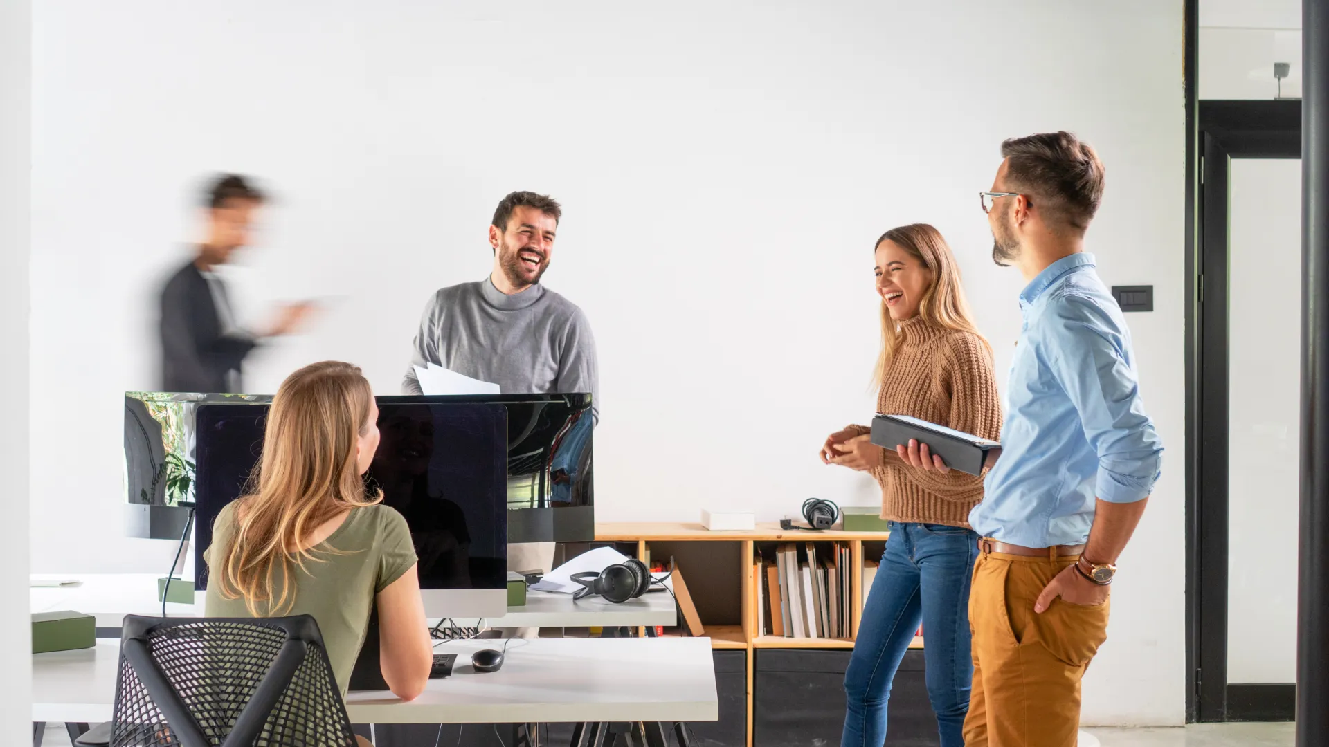 Young professionals casually discussing and laughing in a modern office workspace with computers and notebooks.
