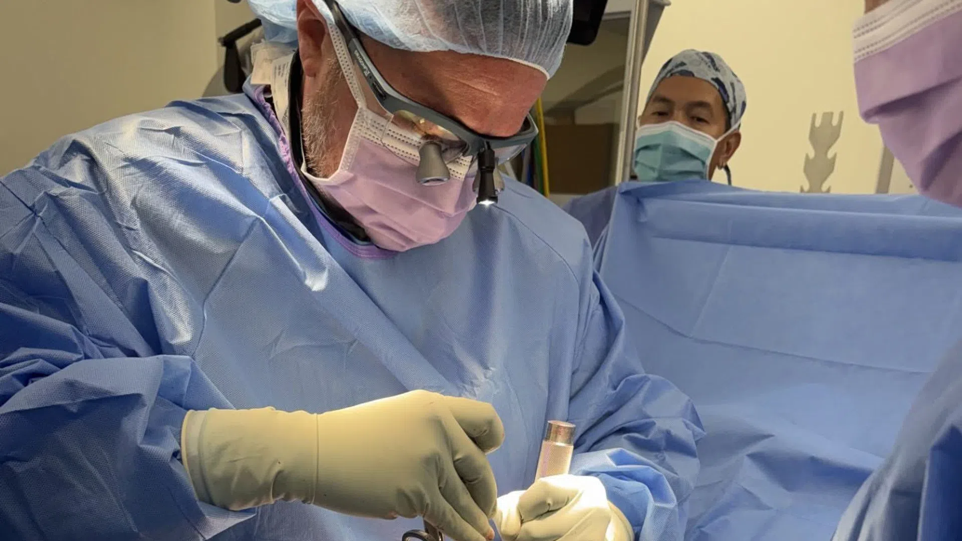 Surgeon wearing magnifying glasses and protective gear performing surgery in an operating room.