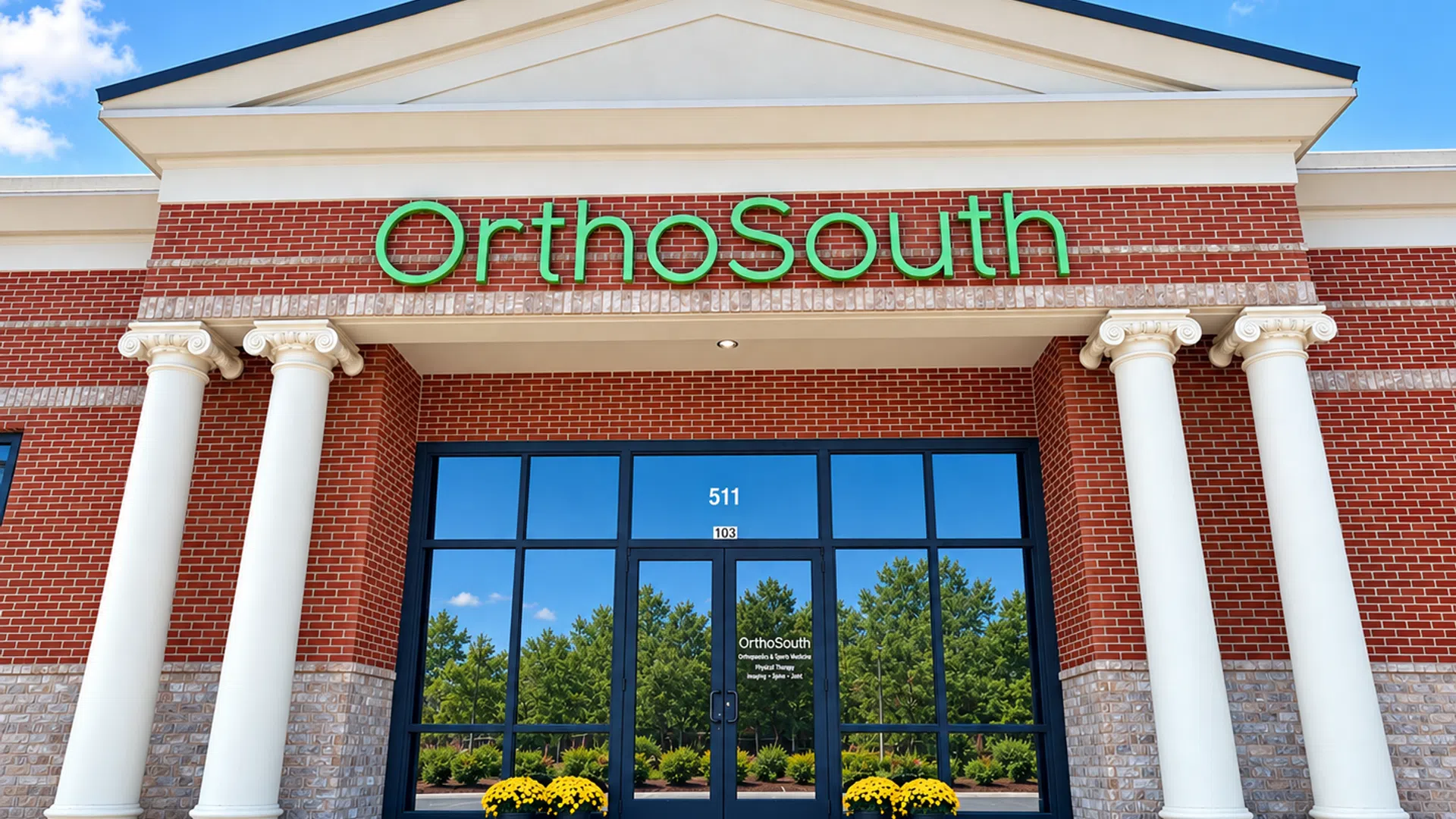 Front entrance of OrthoSouth medical building with columns, red brick, green sign, and glass doors reflecting trees.