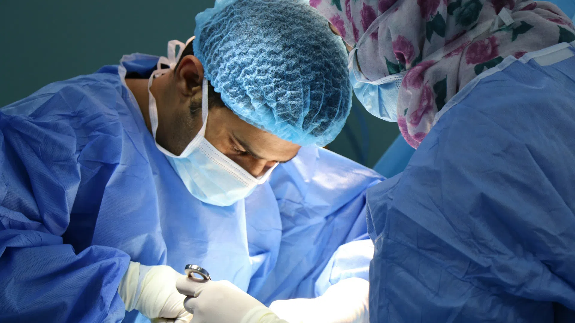 Surgeons in blue scrubs and masks performing a delicate operation in a bright operating room.