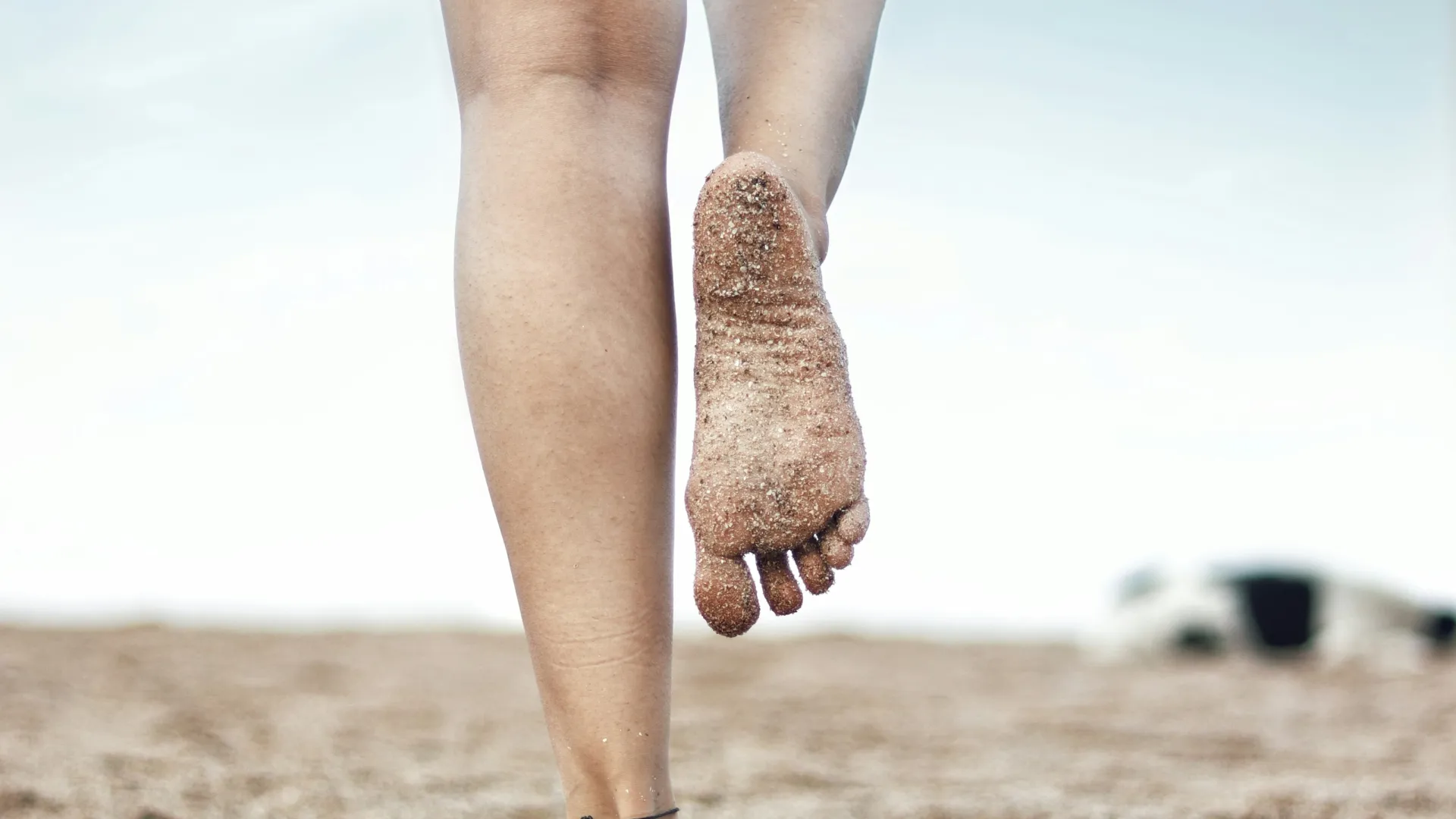 Close-up of a person walking barefoot on sandy beach with one foot lifted showing sand-covered sole.