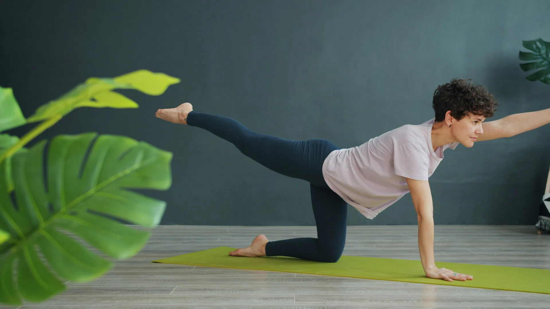Woman practicing yoga balancing on one hand and leg on green mat indoors with plants in the background