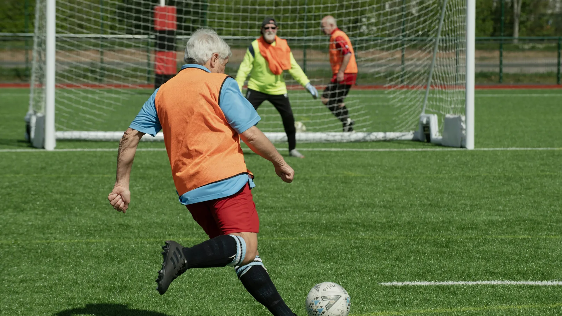 Senior man in orange vest kicking soccer ball toward goal with goalkeeper and defender in background on green field