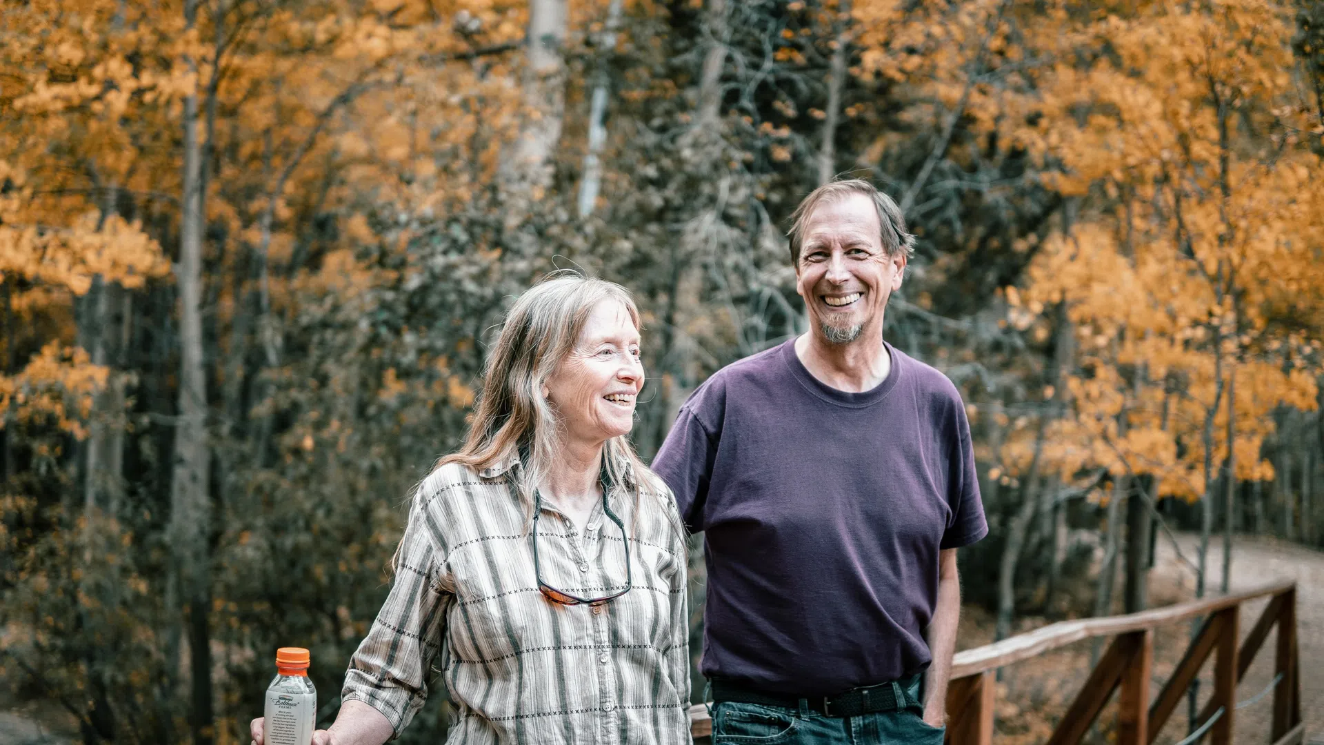 Smiling middle-aged couple enjoys autumn walk on wooden bridge surrounded by fall foliage in warm tones