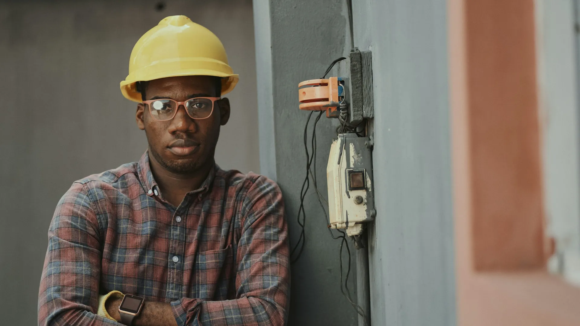 Man in a yellow hard hat and glasses standing next to electrical equipment on a construction site.