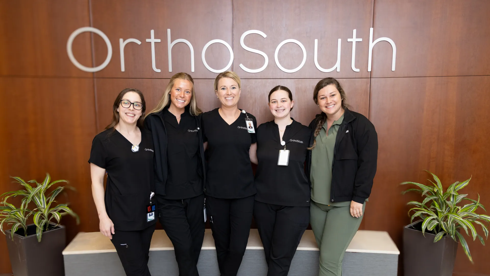 Five healthcare professionals smiling in front of OrthoSouth logo, modern setting with plants and bench.