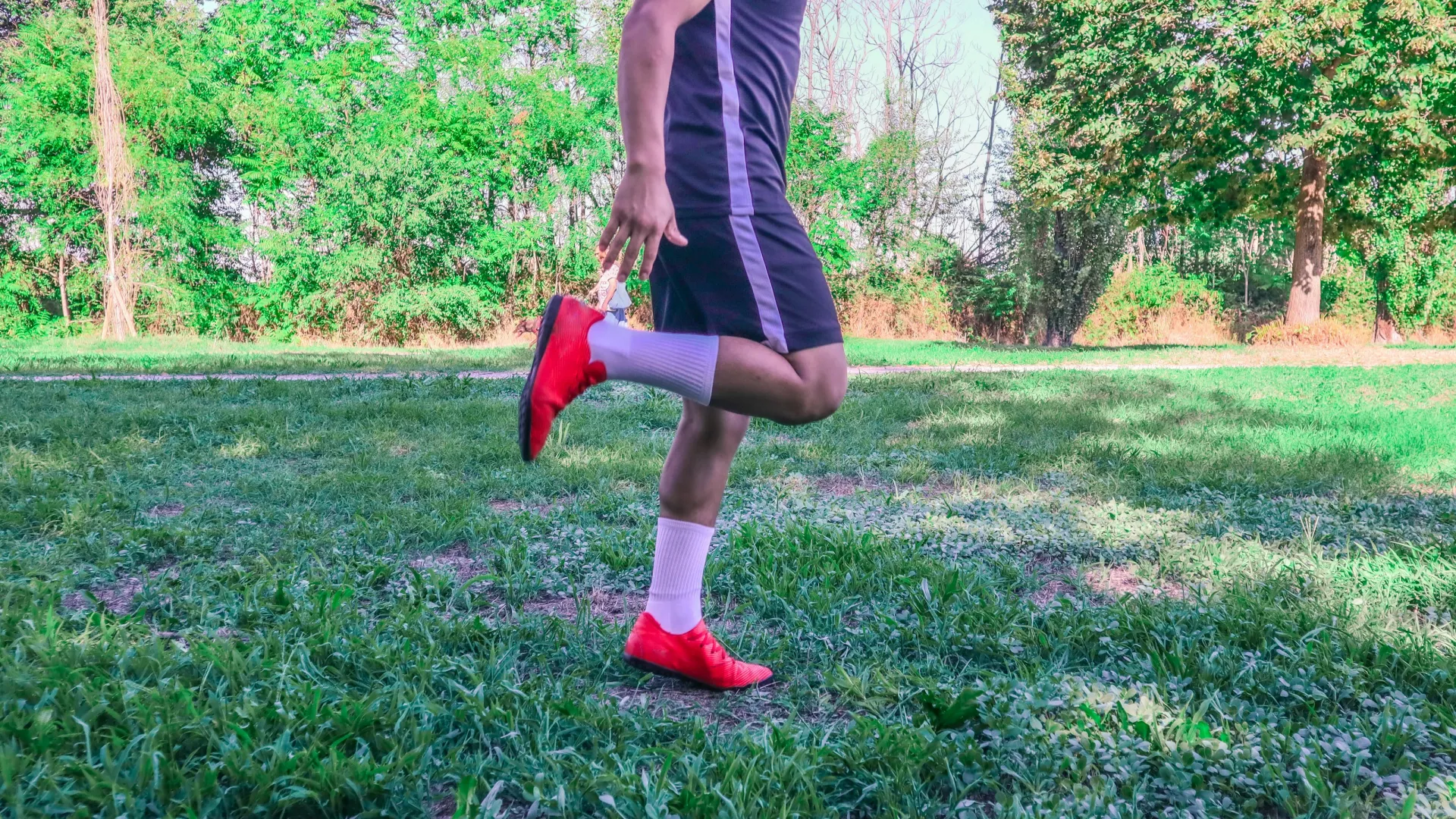 Man in sportswear and red shoes stretching his leg in a green park with trees and grass in the background