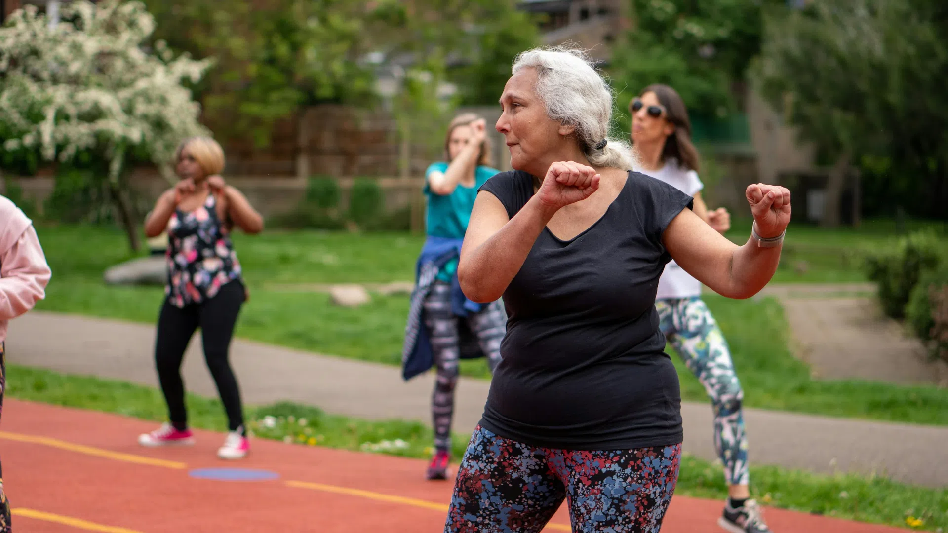 Mature woman in floral leggings doing outdoor group exercise with others on a red running track in park