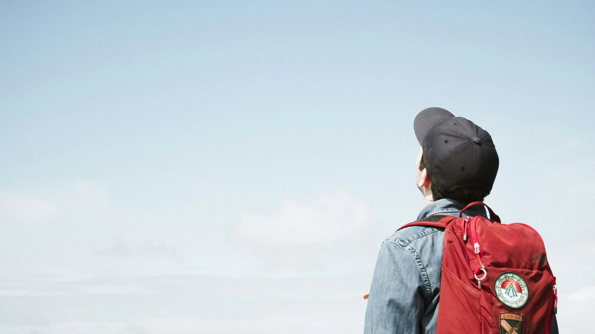 Person wearing a red backpack and black cap looks up at clear blue sky outdoors.