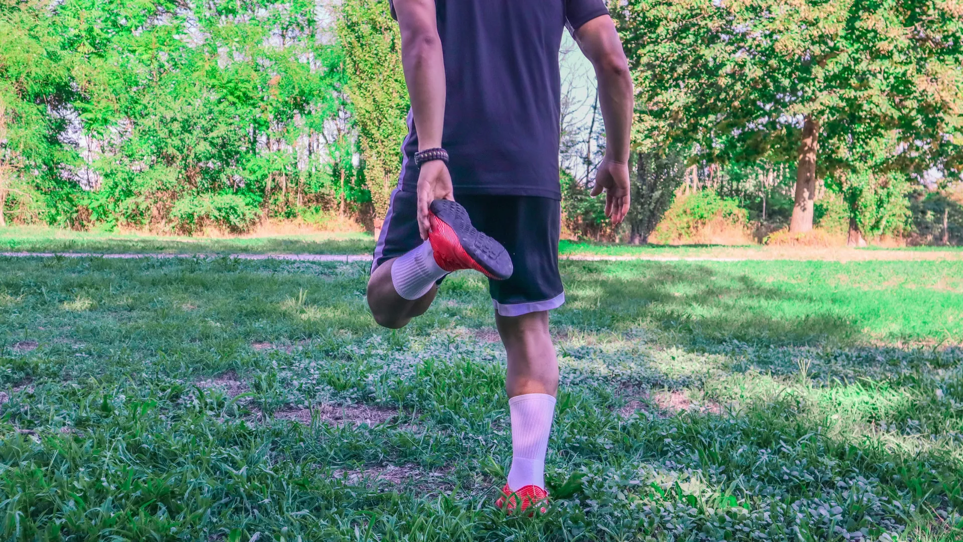 Man stretching outdoors on grass, holding foot behind him, preparing for a run in sunny park.