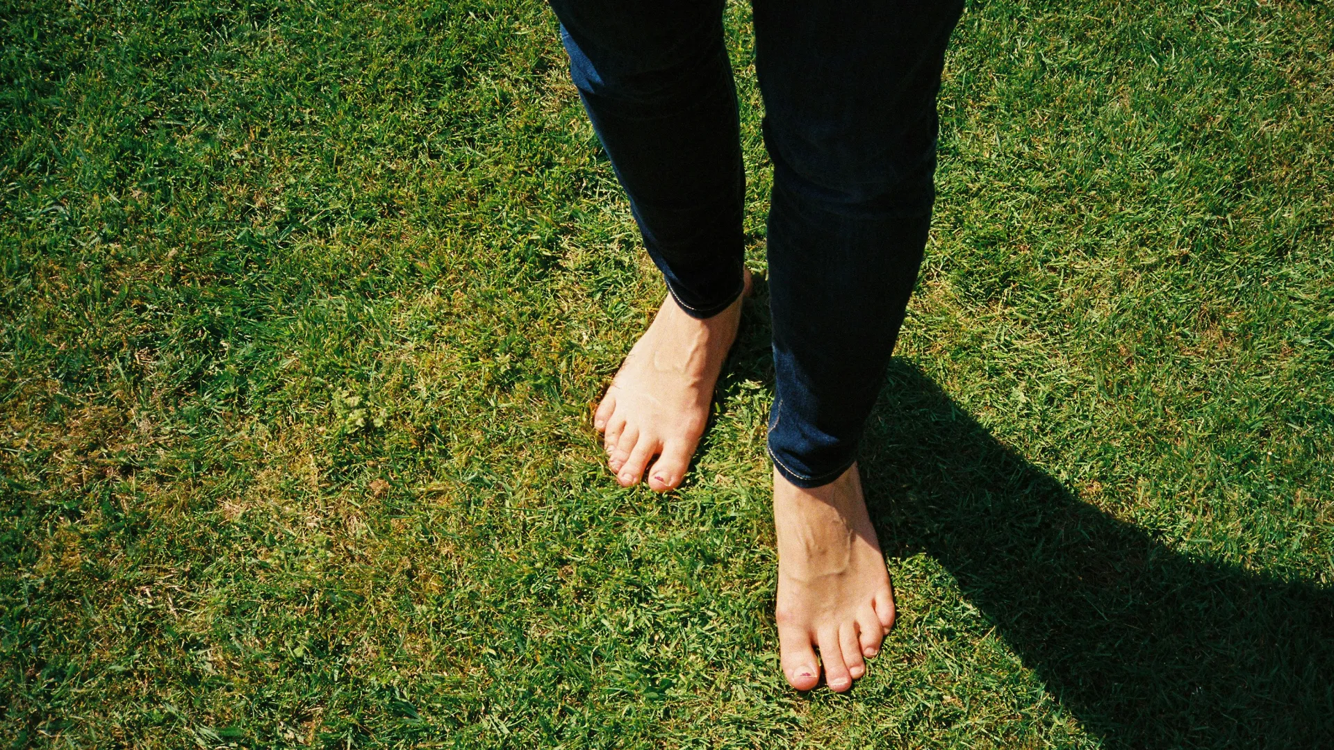Person wearing dark jeans walking barefoot on green grass casting a shadow in daylight