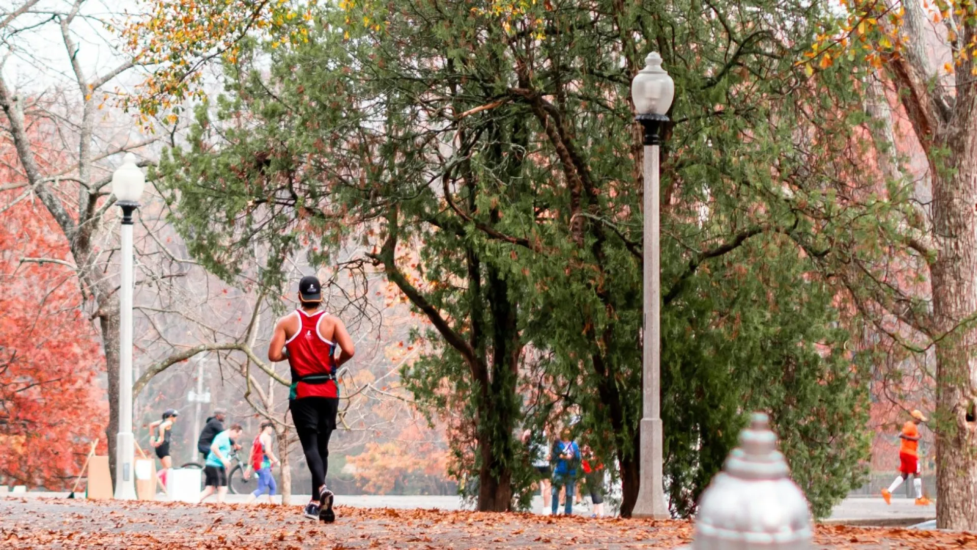 Runner in a red shirt jogging on a leaf-covered park path with fall-colored trees and lampposts around.