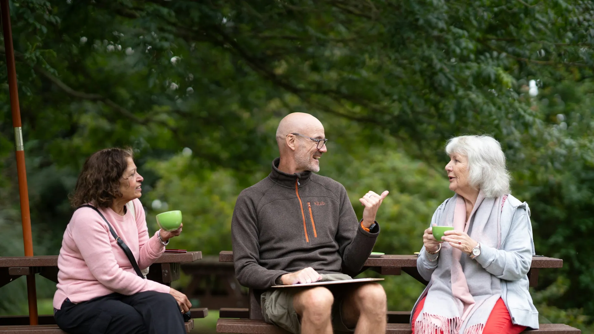 Three older adults sitting on benches outdoors, enjoying drinks and engaging in lively conversation in a park setting.