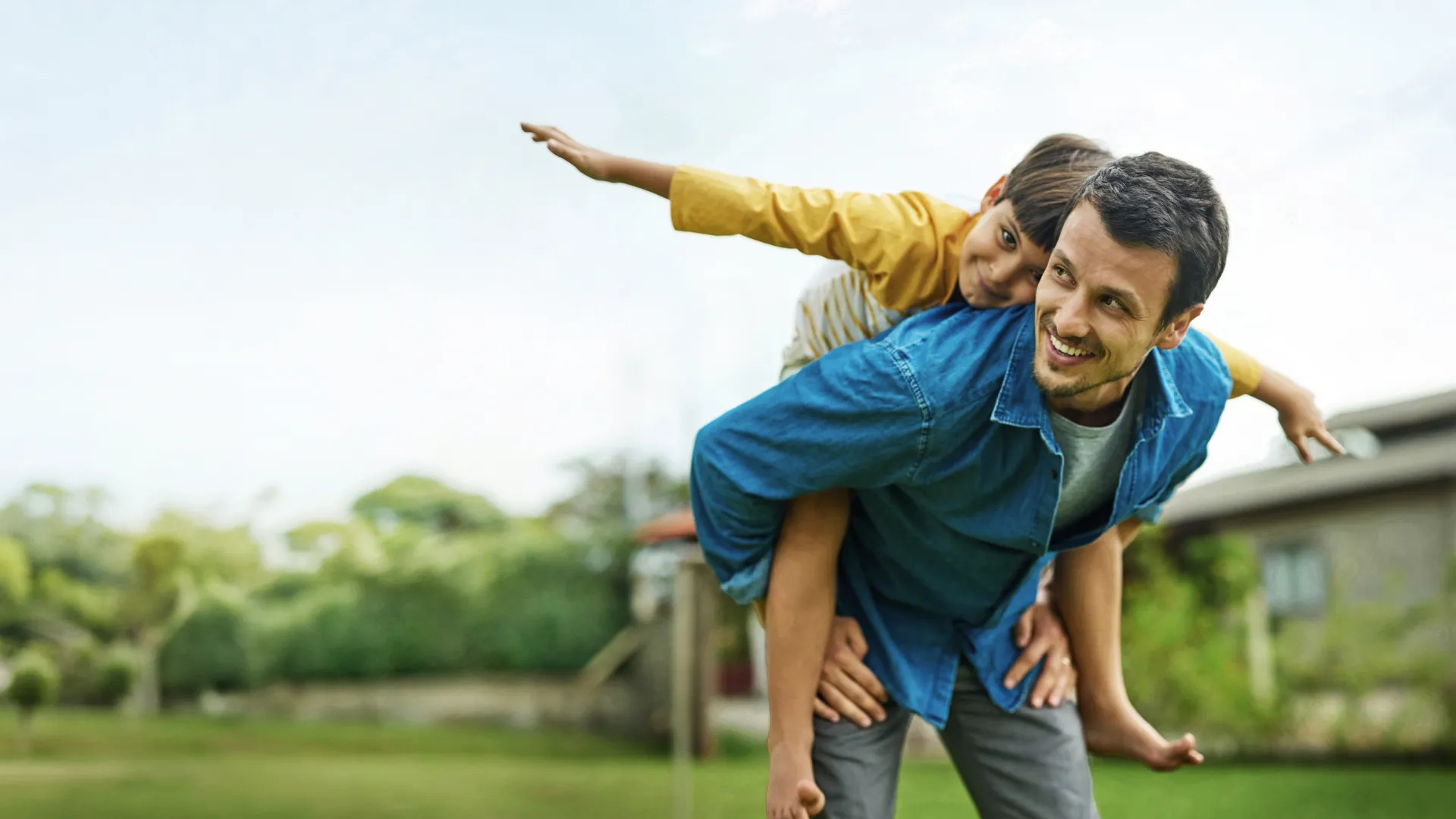 Father giving piggyback ride to son outdoors with greenery and clear sky in background
