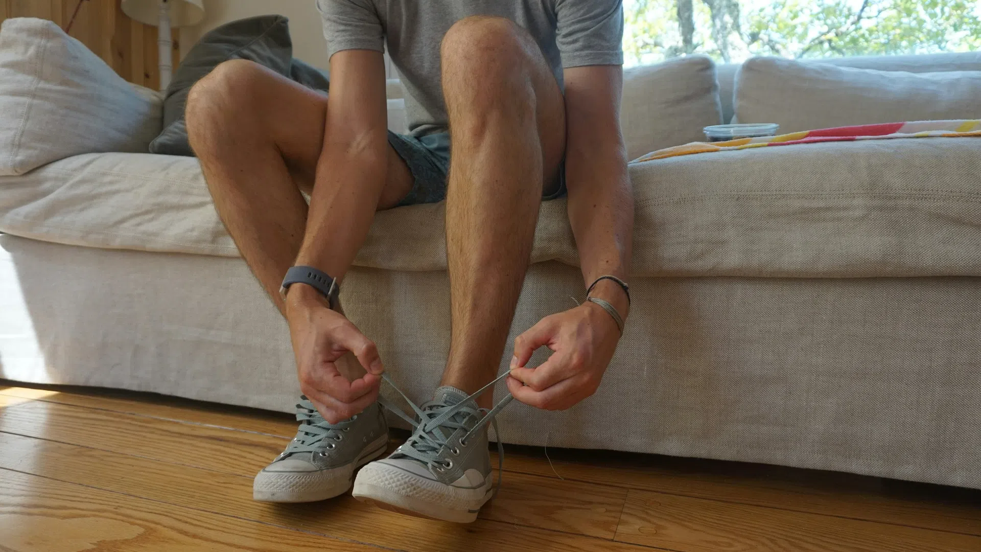 Person sitting on a couch tying shoelaces on light gray sneakers in a sunlit room with wooden floor.