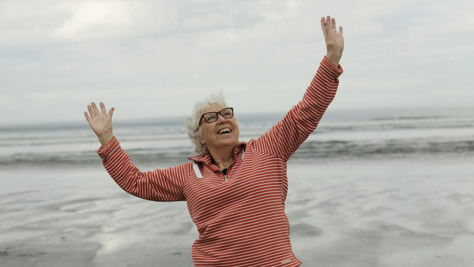 Smiling elderly woman in red striped shirt joyfully raising hands on a cloudy beach with ocean in the background