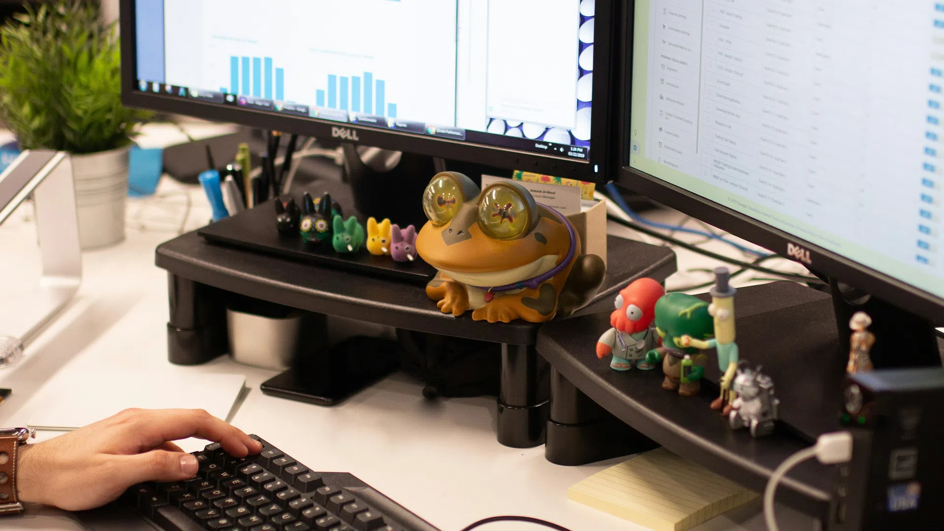 Person working on dual Dell monitors with charts and data, surrounded by desk toys and office equipment.
