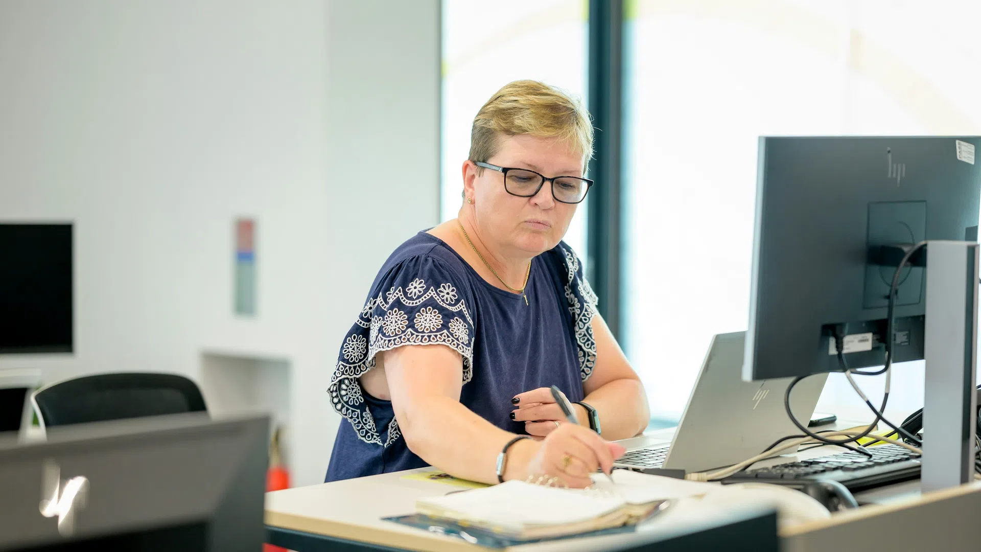 Woman in glasses working at a desk with computer writing in a notebook in a bright modern office.