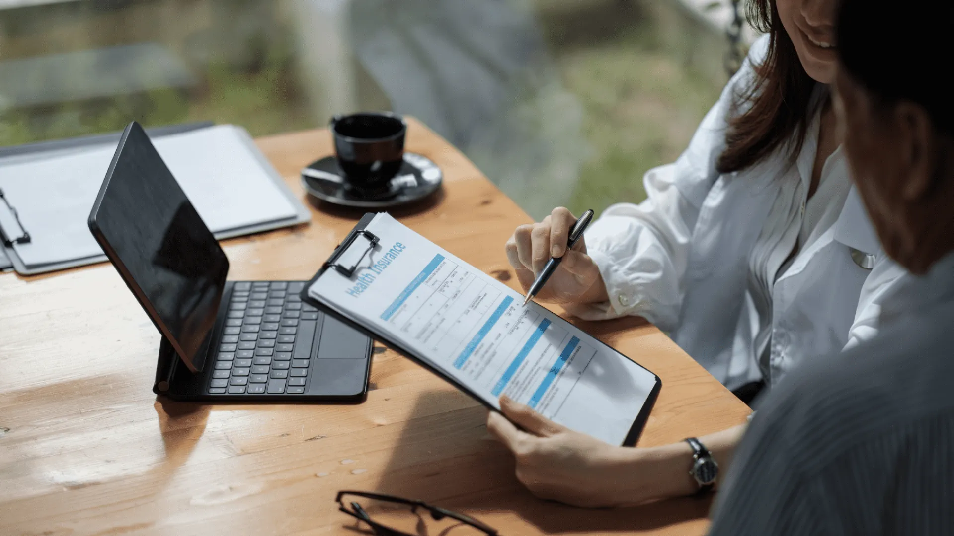 Two people reviewing a health insurance form on a clipboard at a wooden desk with tablet and coffee cup