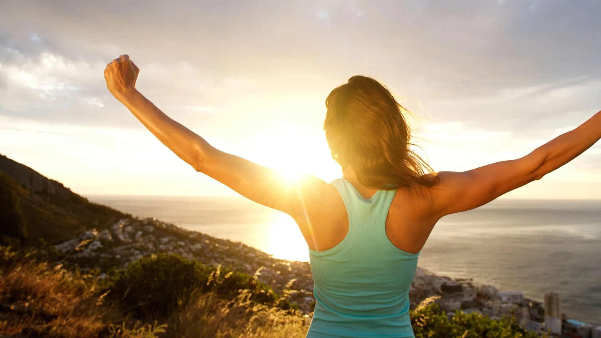 Woman in athletic wear raising arms joyfully at sunrise overlooking ocean and hills.