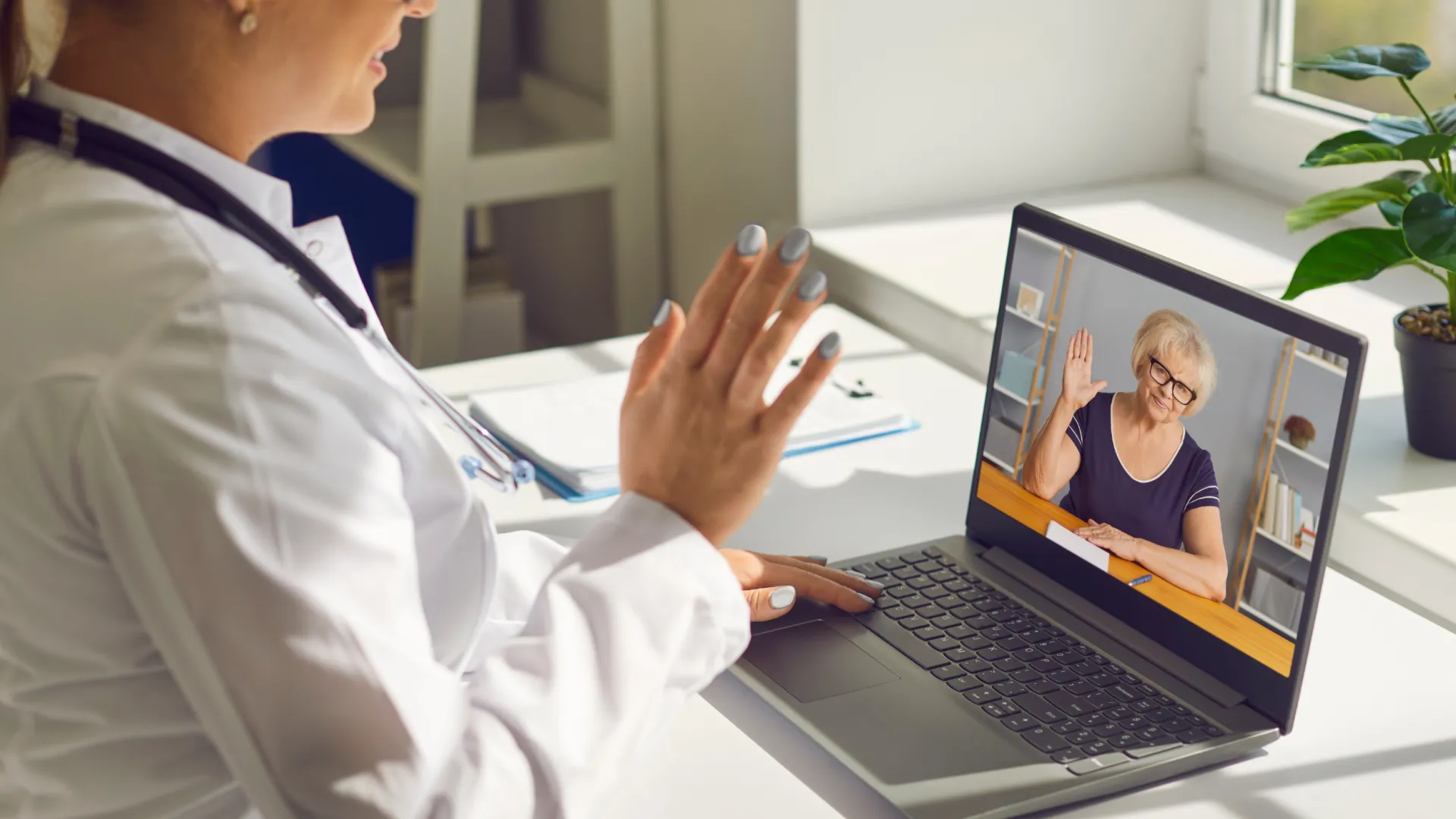 Doctor conducting a video consultation with a senior woman waving on a laptop screen.