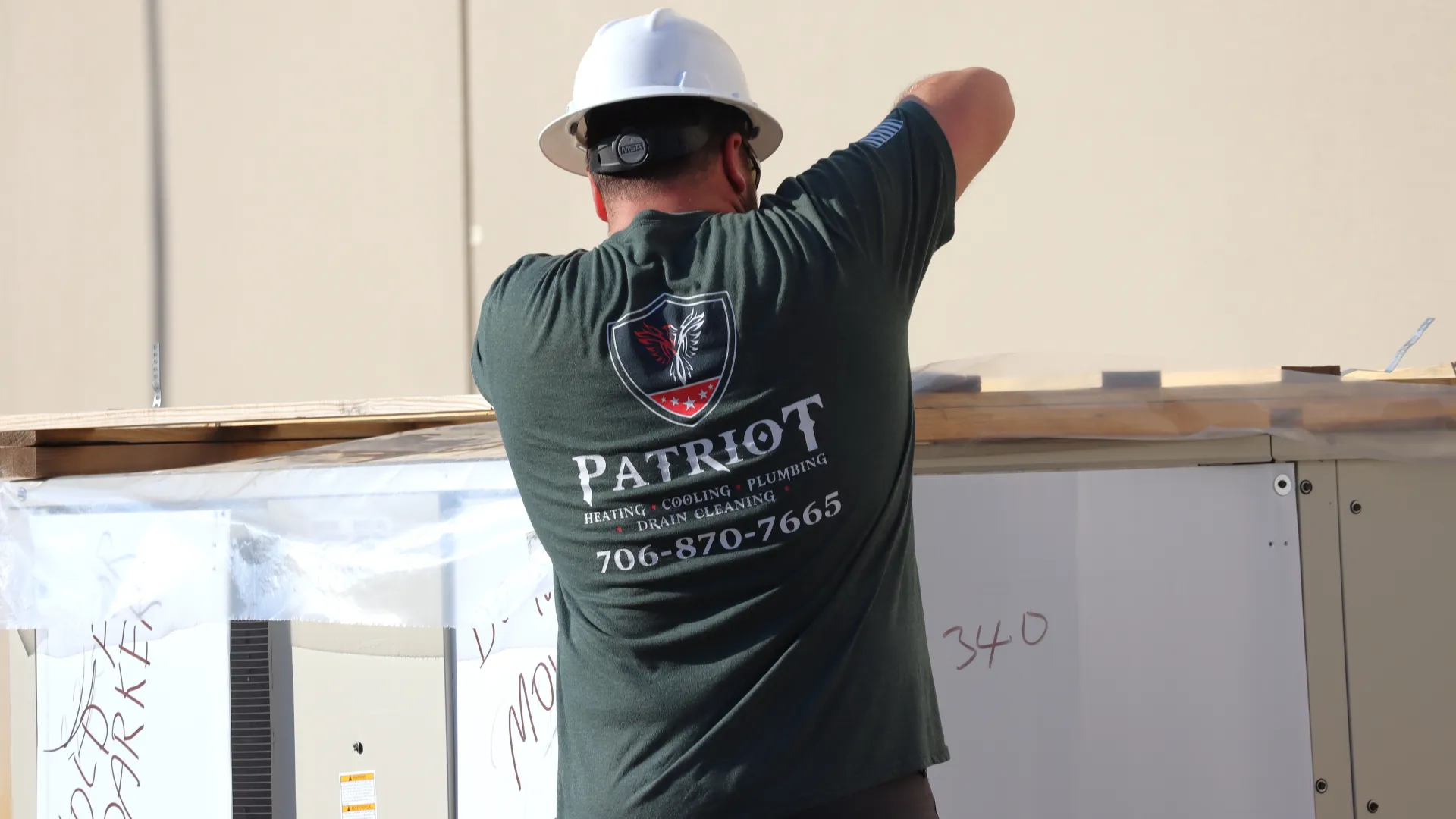 Worker wearing Patriot service company shirt and white hard hat assembling wooden structure outdoors.