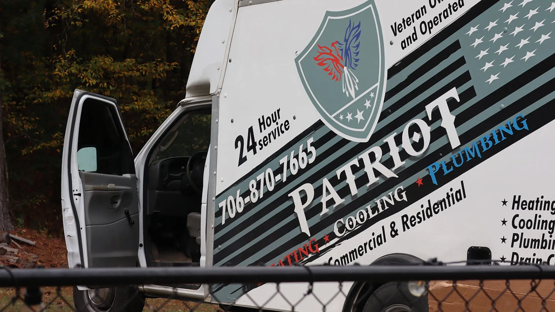 Patriot Heating, Cooling, and Plumbing service van with open door parked near trees and fence.