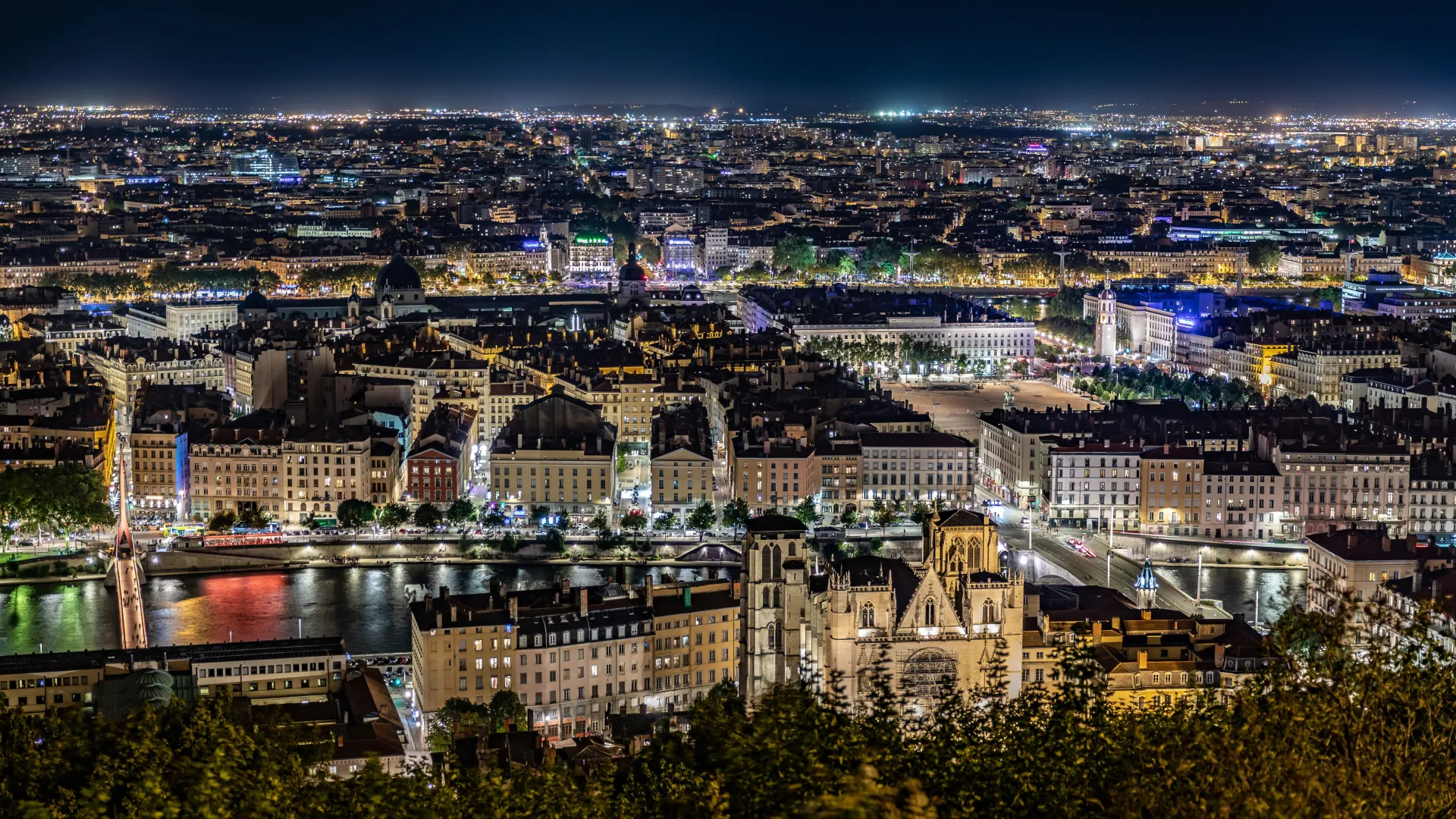 Night view of a European city with illuminated buildings, bridges, river, and streets bustling with lights.