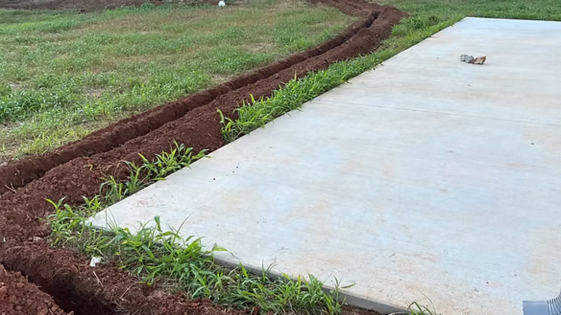 Freshly dug narrow trenches curving around a concrete slab near a building with grass and soil surrounding.
