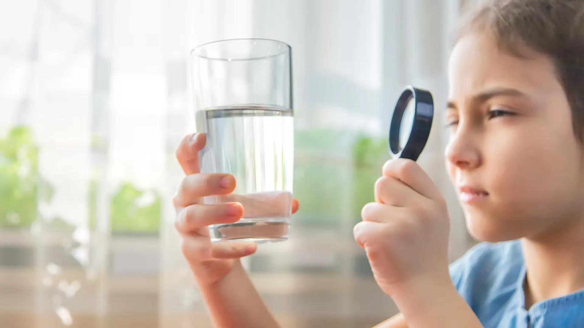 Young girl examining a clear glass of water closely using a magnifying glass indoors.