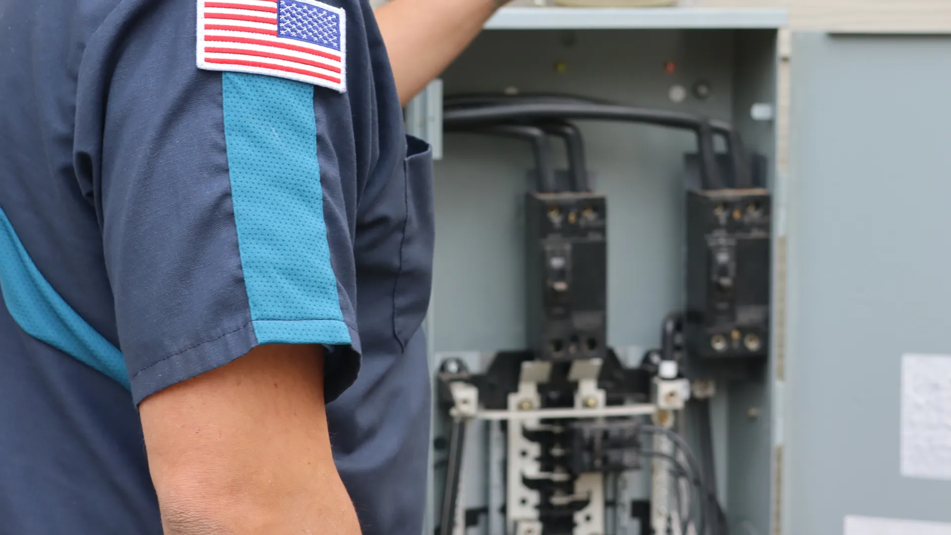 Electrician with US flag patch working on an outdoor electrical panel or circuit breaker box.