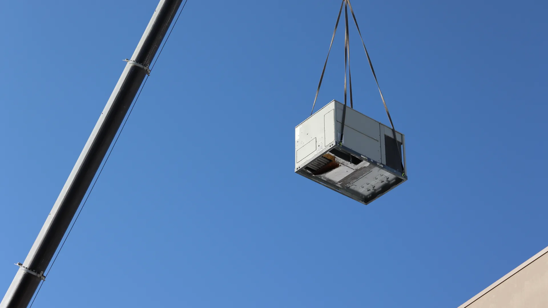 Crane lifting a large HVAC unit against a clear blue sky above a building corner.