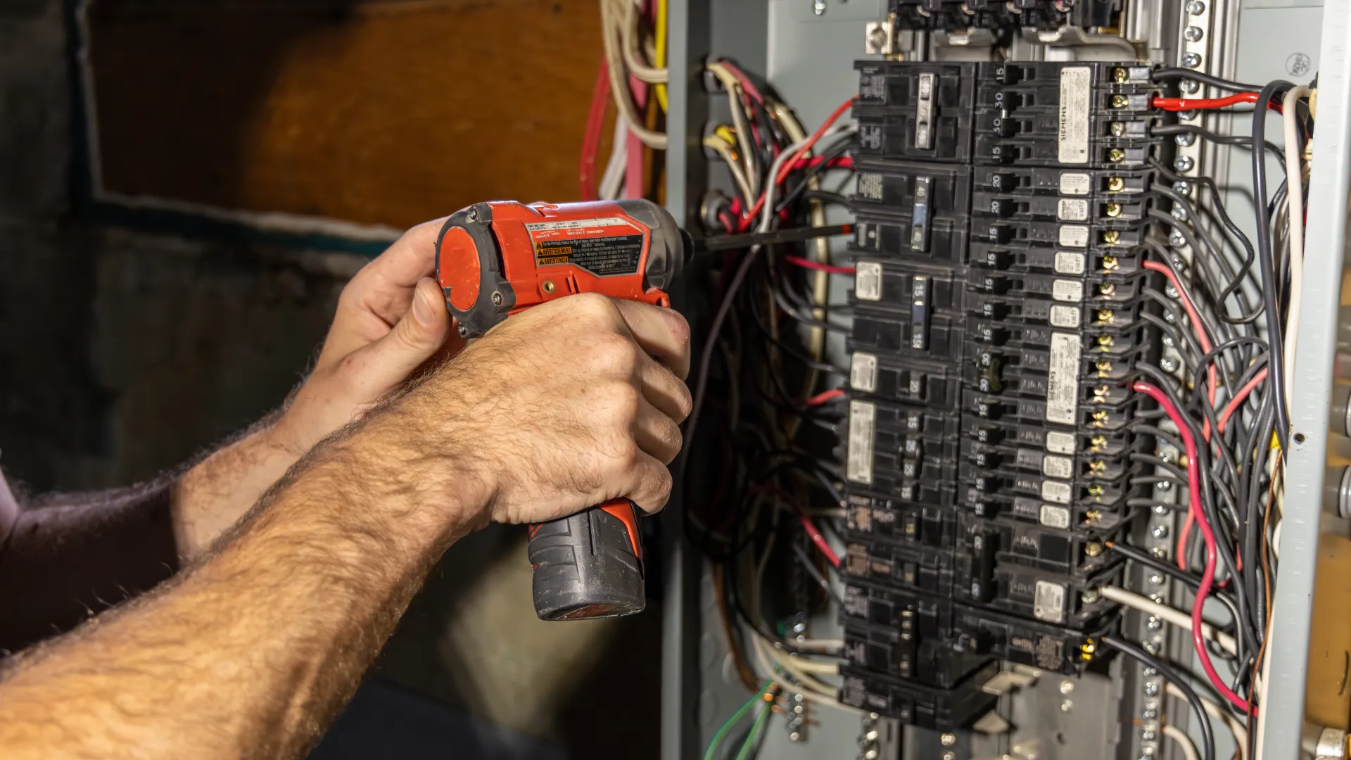 Electrician using a cordless drill to work on a circuit breaker panel with multiple wires and breakers.