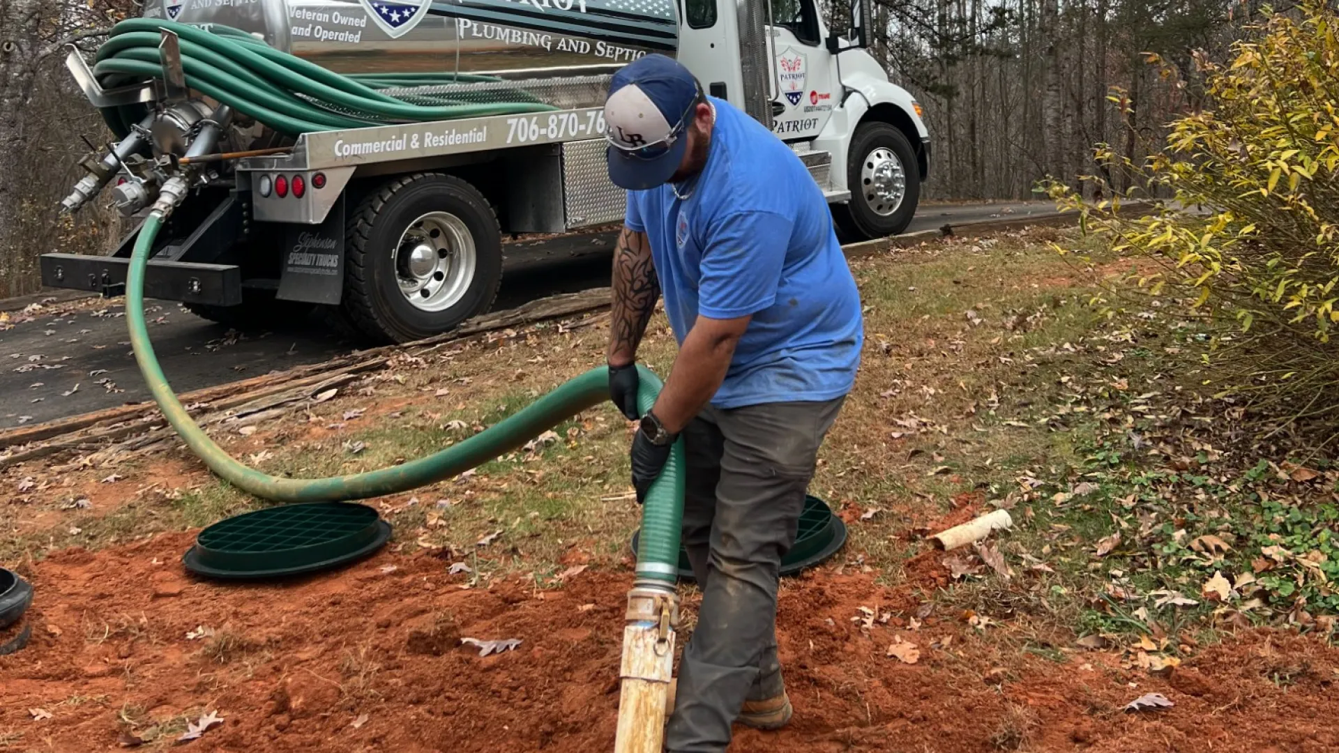 Worker in blue shirt using hose connected to Patriot plumbing truck near open manhole on red dirt ground.