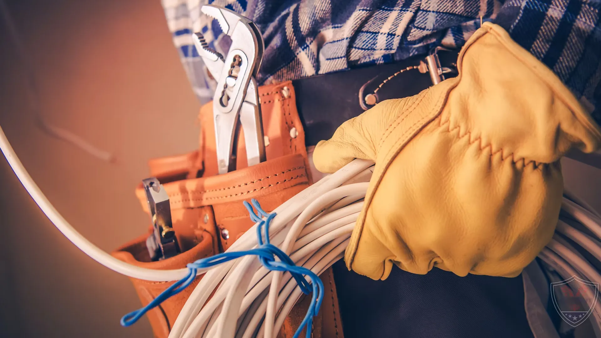 Electrician wearing yellow gloves holding coiled wires with a tool belt containing pliers and screwdrivers.