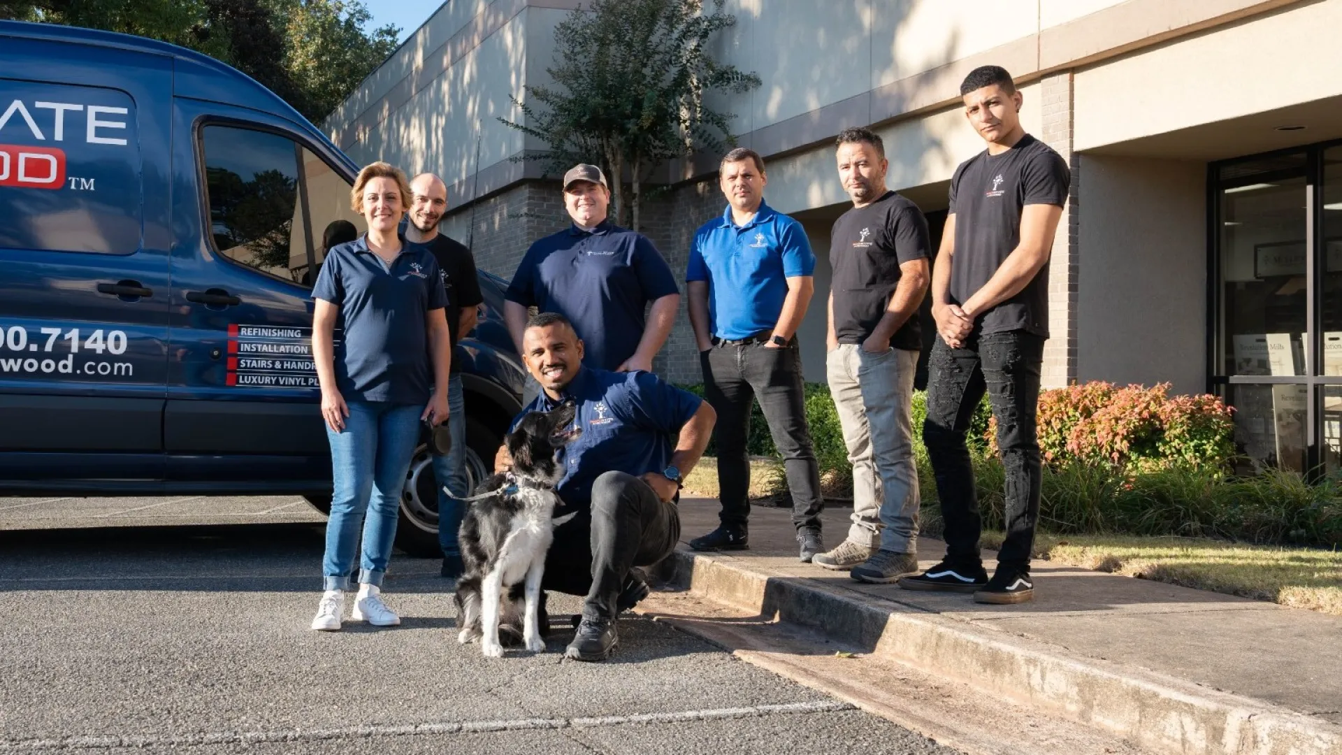 Team of seven workers with a dog posing outside a service van and building on a sunny day