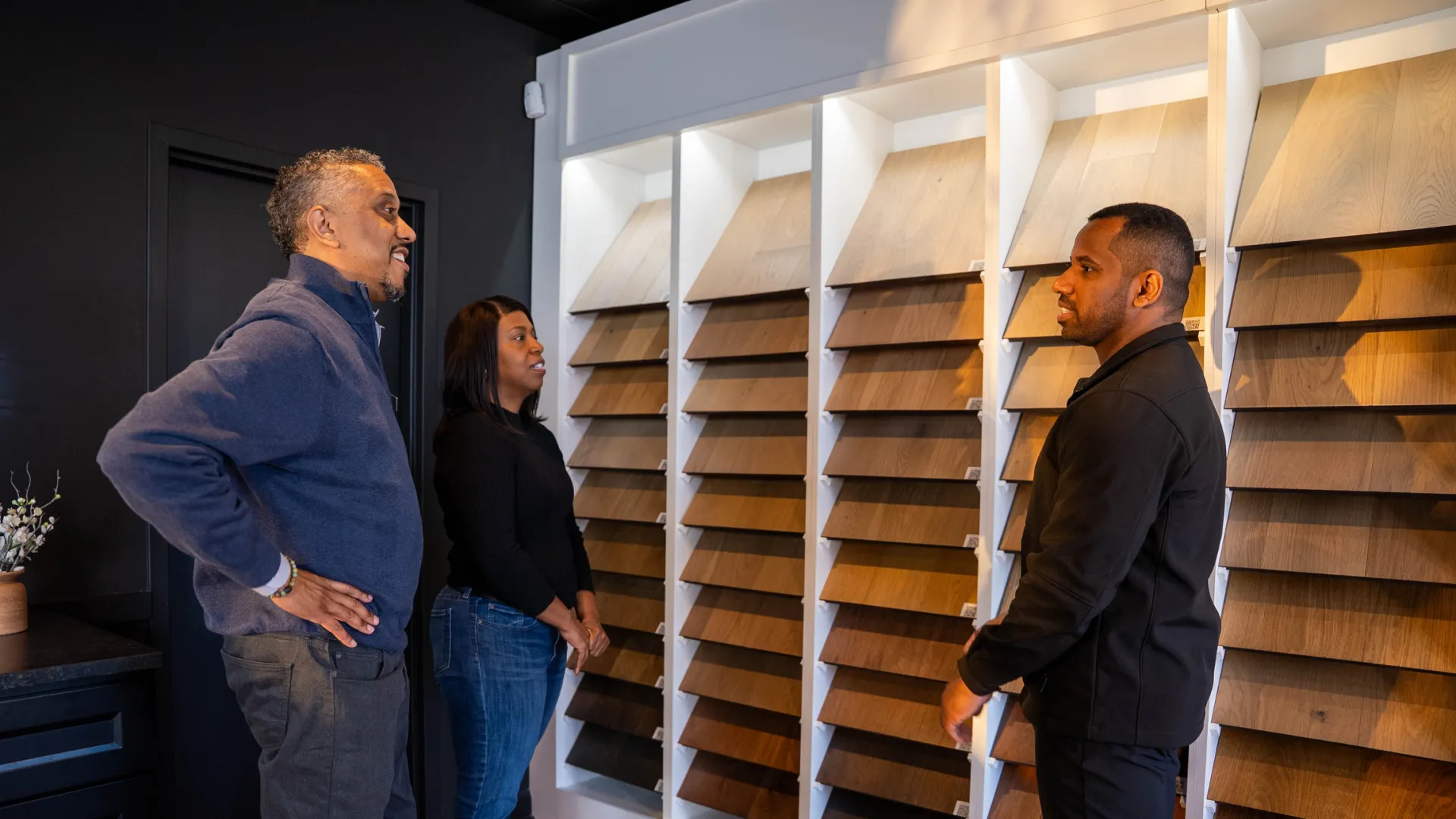 Couple consulting with a salesperson about various wooden flooring samples displayed on wall panels.