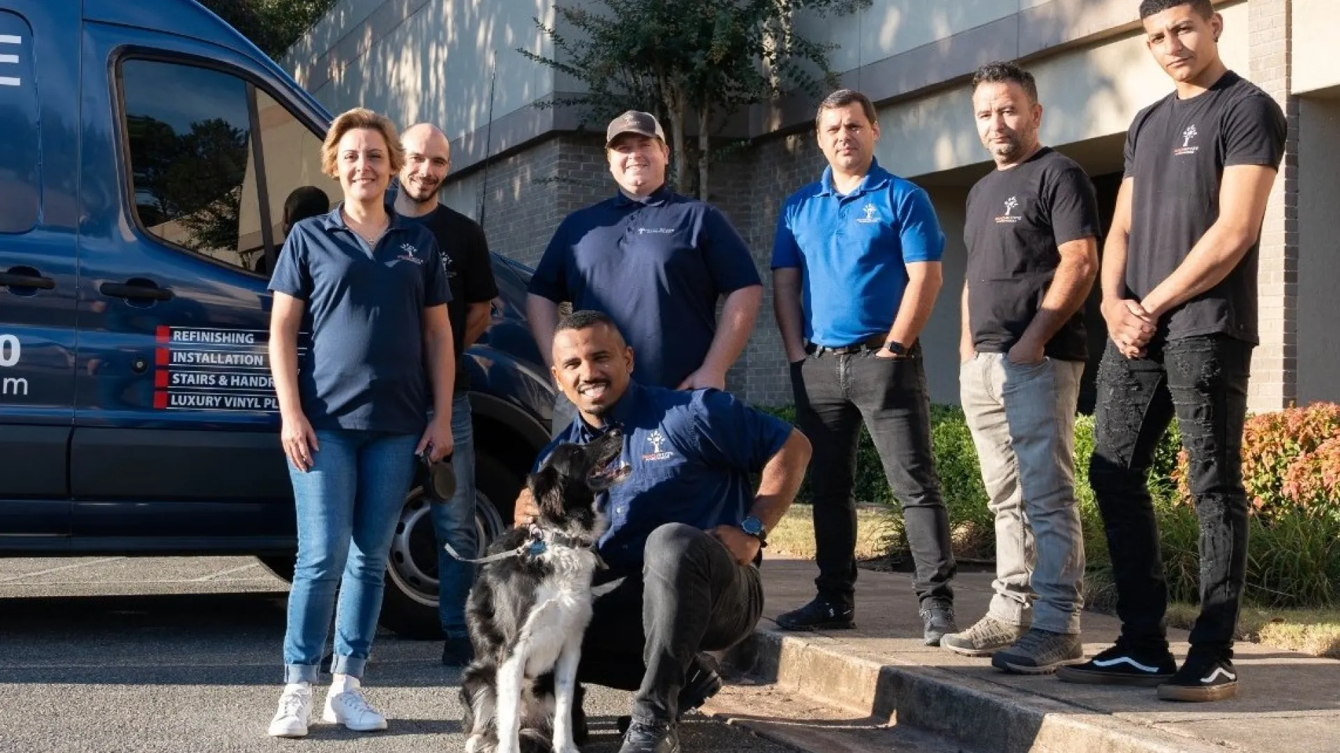 Group of seven workers standing outside a building near a company van, one kneeling with a black and white dog.